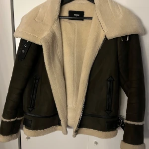 Brun pilotjacka med fuskpäls från Bikbok - Säljer en brun pilotjacka med beige fuskpälsfoder och stora slag. Jackan har dragkedja, bältesdetaljer och svarta skinnliknande detaljer vid fickor och ärmslut. Perfekt nu för våren 