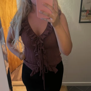 Vinröd ribbad topp med volang och knyt - Säljer en vinröd ribbad topp med v-ringning, volangdetaljer längs kanten och knyt framtill. Perfekt för dig som gillar feminina detaljer och en trendig look.