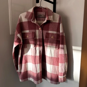 Bianca by Nelly jacka - Säljer en jacka från Bianca by Nelly i rosa och vitt rutigt mönster. Skjortan har två stora bröstfickor med knappar och klassisk krage. Perfekt att slänga över en t-shirt för en avslappnad look.