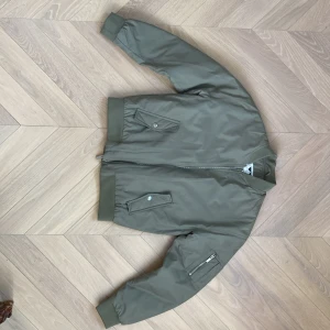 Olivgrön bomberjacka från Junkyard - Säljer en olivgrön bomberjacka från Junkyard med orange quiltat foder. Jackan har dragkedja, fickor med lock och ärmlång ficka med dragkedja. Klassisk bombermodell med ribbade muddar vid ärmslut och midja.