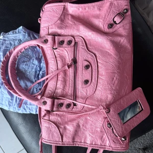 Rosa axelväska med spegeldetalj - Säljer en snygg rosa axelväska i syntetmaterial med coola nitar, dragkedja framtill och spegeldetalj. Väskan har dubbla handtag och dekorativa remmar. Perfekt för dig som vill sticka ut med färg och stil.