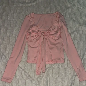 Rosa långärmad topp med knyt - Säljer en söt rosa långärmad topp med v-ringning och knyt framtill. Toppen är croppad och har en mjuk, stretchig känsla. Perfekt för att skapa en trendig look.