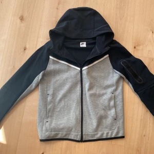 Nike Tech Fleece Hoodie - Nike hoodie med dragkedja i grått och svart. Jackan har huva, långa ärmar och två fickor framtill. Snygg sportig look med svarta detaljer på axlar och ärmar samt en vit linje över bröstet.