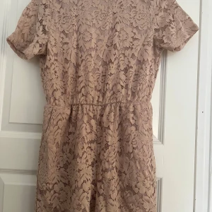 Beige spetsbyxdress från NA-KD - Säljer en superfin beige byxdress med korta ben från NA-KD i storlek 36. Klänningen har korta ärmar, blommigt spetsmönster och en snygg öppen detalj i ryggen. Perfekt för vår och sommar när du vill känna dig extra söt.