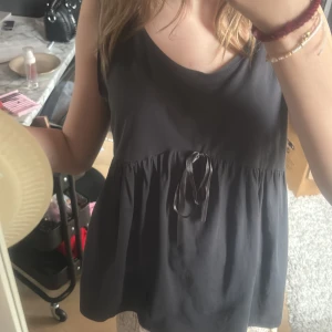 Svart ärmlös topp  från me&i - Svart ärmlös topp från me&i med rund halsringning och ett dekorativt band i midjan. Klänningen har en avslappnad passform och är tillverkad i mjukt material, perfekt för varma dagar.