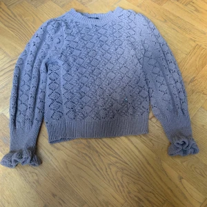 Ljuslila stickad tröja från Lindex - Superfin brun stickad tröja från Lindex med vackert hålmönster och volangdetaljer vid ärmsluten. Tröjan har lång ärm och ribbade muddar. Perfekt för lager-på-lager under kyliga dagar.
