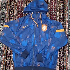 Blå vindjacka från Nike med huva och orange detaljer - Säljer en blå vindjacka från Nike med huva, dragkedja och orange detaljer. Jackan har ett mönster i olika blå nyanser, orange meshfoder och ribbade muddar. Nederdelen av ärmarna har en orange rand och bröstet pryds av ett vitt lejonemblem.