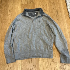 Grå halvzip tröja från Polo Ralph Lauren - Säljer en stilren grå halvzip tröja från Polo Ralph Lauren. Tröjan har hög krage, dragkedja vid halsen och diskret broderad logga på bröstet. Perfekt för en avslappnad men snygg look.
