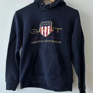Mörkblå hoodie från Gant - Säljer en helt ny klassisk mörkblå hoodie från Gant med broderad logga och texten 'American Sportswear' framtill. Tröjan har huva, magficka och ribbade muddar. Perfekt för en sportig och avslappnad stil.