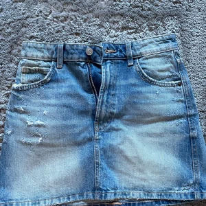 Blå jeanskjol från Zara - Säljer en klassisk blå jeanskjol från Zara i kort modell. Kjolen har slitna detaljer framtill, femficksdesign och stängs med dragkedja och knapp. Perfekt till sommaren och enkel att matcha med olika toppar.
