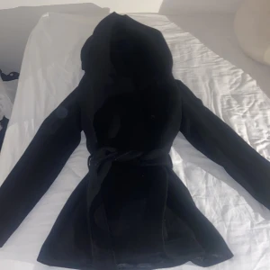 Svart kappa med bälte från Amisu - Säljer en svart kappa från Amisu i storlek 34. Jackan har en stor luva, knappar och bälte i midjan. Är använd 3 gånger och ser ut som ny.