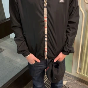 Svart vindjacka från Burberry - Säljer en stilren svart vindjacka från Burberry med dragkedja och klassiskt rutigt mönster på insidan av kragen. Jackan har långa ärmar och en huva för extra skydd mot väder. Perfekt för en casual look.