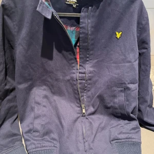 Marinblå vindjacka från Lyle & Scott - Snygg marinblå vindjacka från Lyle & Scott med dragkedja framtill och klassisk gul logga på bröstet. Jackan har ribbade muddar och fickor på sidorna samt rutigt innerfoder för extra stil. Storlek 12-13 år. 200kr. 