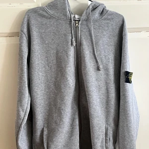 Grå hoodie med dragkedja från Stone Island - Säljer en grå hoodie från Stone Island med dragkedja framtill och huva med snören. Tröjan har långa ärmar och klassisk logotyp på vänster ärm. Perfekt för en avslappnad stil.