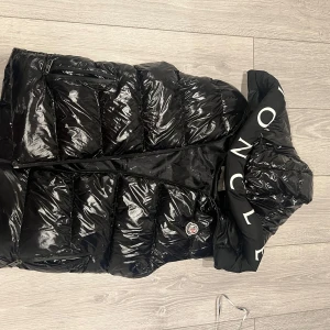 Svart dunväst från Moncler med huva - Säljer en svart, glansig dunväst från Moncler i storlek S. Västen har en stor huva med Moncler-logga i vitt, dragkedja framtill och klassisk quiltad design. Perfekt för lager-på-lager och riktigt snygg till streetwear-looken.