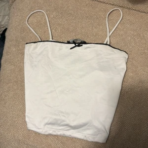 Vit linnetopp med svarta detaljer FB Sister - Supersöt vit linnetopp från FB Sister med tunna axelband och svarta spetsdetaljer längs överkanten. Toppen har en liten svart rosett framtill som ger en gullig touch. Perfekt att styla med jeans eller kjol för en trendig look.