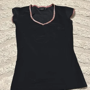 Svart topp med rosa kant från Casall - Svart kortärmad topp från Casall med v-ringning och rosa mönstrad kant runt hals och ärmslut. Snygg och sportig passform, perfekt för dig som gillar en enkel men ändå detaljrik look. Materialet känns lätt och smidigt.