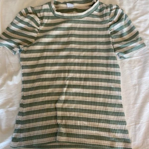 Randig ribbad t-shirt från Lindex - Superfin ribbad t-shirt från Lindex med gröna och vita ränder samt tunna silvertrådar som ger lite extra bling. Kortärmad och stretchig, perfekt till jeans eller shorts. Skön passform och mjukt material.