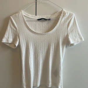 Vit ribbad topp från Vero Moda - Supersöt vit ribbad topp från Vero Moda med korta ärmar och rundad halsringning med liten volangkant. Tunt, mjukt material i bomull som känns skönt mot huden. Perfekt till jeans eller kjol för en fräsch look.