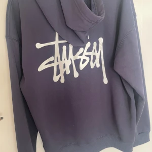 Marinblå stussy hoddie - Säljer en mörklila hoodie från Stüssy med deras ikoniska vita logga tryckt stort på ryggen och mindre på bröstet. Tröjan har huva med snörning och känguruficka framtill. Perfekt för dig som gillar streetwear och vill sticka ut.