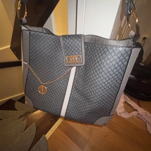 Grå axelväska från River Island - Snygg grå axelväska från River Island med diskret mönster, guldfärgade detaljer och kedja framtill. Väskan har bred axelrem med stora ringar och praktiskt innerfack med dragkedja. Perfekt för dig som vill ha en stilren och rymlig väska.