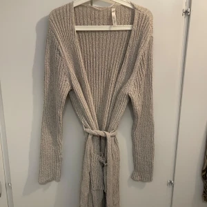 Beige ribbad kofta med knytband - Lång, ribbstickad beige kofta från H&M med knytband i midjan och två stora fickor framtill. Perfekt att slänga på sig över en t-shirt eller linne. Koftan har långa ärmar och en relaxed fit som ger en chill vibe.