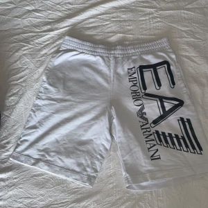 Vita shorts från Emporio Armani EA7 - Snygga vita shorts från Emporio Armani EA7 med stor svart logga på ena benet och mindre logga bak. De har elastisk midja och är tillverkade i mjuk bomull, perfekta för sommardagar. Klassisk passform och stilren look.