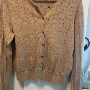 Beige stickad kofta från Kappahl - Snygg beige stickad kofta från Kappahl i storlek S. Koftan har ett genomgående hålmönster, ribbade muddar och knappar framtill. Perfekt att slänga över en topp för en chill och stilren look.