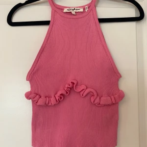 Rosa ribbad linnetopp med volang - Supersöt rosa ribbad linnetopp med volangdetalj över magen. Toppen är ärmlös och har en hög halsringning som ger en trendig look. Perfekt för att lyfta vilken outfit som helst med sin färg och coola design.