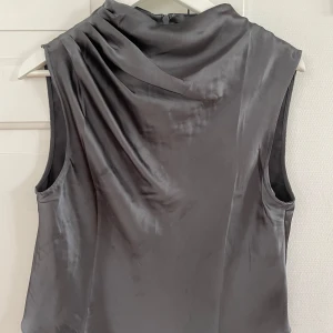 Grå/silver satinblus från H&M - Snygg grå/silver blus i satin från H&M med draperad detalj vid axeln och hög hals. Ärmlös modell med diskret dragkedja bak🩶🪩