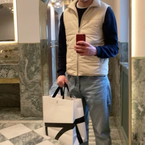 Beige dunväst från These Glory Days nypris 1399 - Stilren beige dunväst från These Glory Days med quiltad design och hög ribbad krage. Västen har två fickor med dragkedja och hel dragkedja framtill. Perfekt att slänga över en hoodie eller tröja för en clean look.