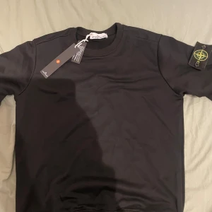 Svart sweatshirt från Stone Island M - Svart sweatshirt från Stone Island i klassisk modell med rund hals och ribbade muddar. Tröjan har den ikoniska Stone Island-loggan på vänster ärm. Tillverkad i mjuk bomull och passar perfekt till streetwear-stilen.