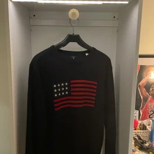 Svart stickad tröja från Gant - Svart stickad tröja från Gant med lång ärm och rund hals. På bröstet finns en broderad amerikansk flagga i rött och vitt. Perfekt för dig som gillar klassisk och stilren design med en sportig touch. Storlek L kan passa m också. Knappt använd