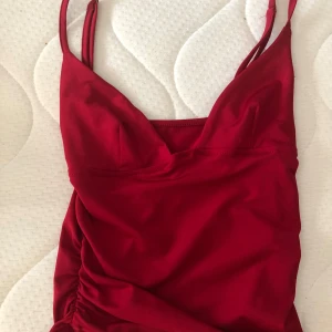 Röd kort klänning med smala axelband - Snygg röd kortklänning med smala axelband och v-ringad urringning. Klänningen har en figurnära passform och lätt rynkade detaljer i sidan. Perfekt för dig som vill sticka ut med en enkel men stilren look.