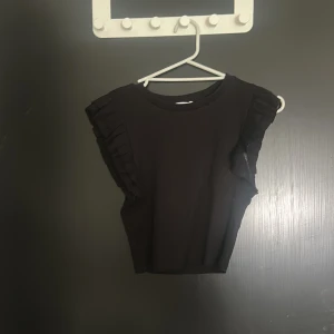 Svart croppad topp med volang från Zara - Säljer en svart croppad topp från Zara med volangdetaljer på axlarna. Toppen är ärmlös och har en enkel, stilren design som passar perfekt till jeans eller kjol. Materialet känns mjukt och stretchigt för en bekväm passform. Storleken är M, men jag tycker att den ändå är ganska liten så känns som en S/XS