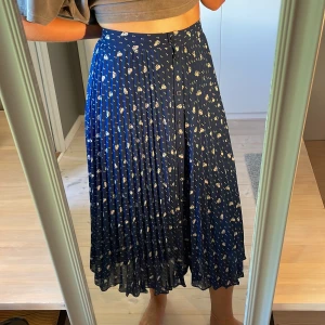 Blommig plisserad midikjol i blått - Supersöt blå långkjol med vita blommor och små prickar. Kjolen är plisserad och har knappar framtill som snygg detalj. Perfekt flow och midjehög passform som ger en riktigt fin siluett. Lätt och luftig, passar perfekt till vår och sommar.