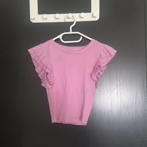 Rosa volangtopp - Supersöt ljuslila/rosa topp från Zara med volangdetaljer på axlarna. Toppen är ribbad och har en rund halsringning samt korta ärmar med dubbla volanger. Perfekt för dig som vill ha en feminin och trendig look. 