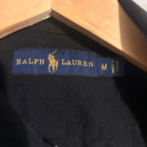 Svart skjorta från Ralph Lauren M - Klassisk svart skjorta från Ralph Lauren i bomull med knappar framtill och röd broderad logga på bröstet. Skjortan har lång ärm och button down-krage, perfekt för en clean och stilren look. Priset är inte hugget i sten så kom med bud
