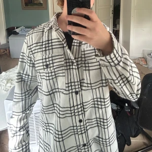 Vit och svart rutig overshirt - Säljer en snygg vit overshirt med svart rutigt mönster. Skjortan har två bröstfickor med knappar, klassisk krage och långa ärmar. Perfekt att slänga över en t-shirt för en chill look. Materialet känns mjukt och lite tjockare.