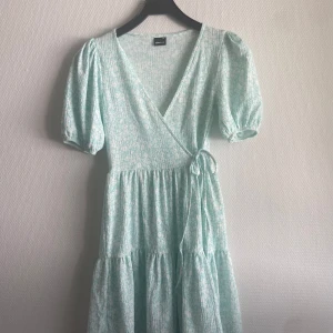 Ljusblå omlottklänning från Gina Tricot - Ljusblå omlottklänning med puffärmar och knyt i midjan. Blommigt mönster och v-ringning.