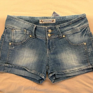 Blå lågmidjade jeansshorts  - Snygga blå låglidjade jeansshorts från DND Fashion med klassisk femficksdesign, dubbla knappar och uppvikta benslut. Shortsen har ljusa slitningar och detaljerade sömmar för en cool look. 