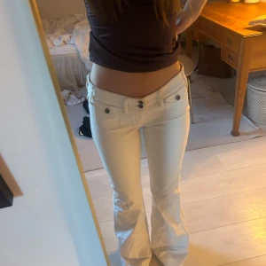 Vita lågmidjade bootcut jeans  - Säljer ett par vita jeans med låg midja och bootcut-ben. Byxorna har dubbla knappar framtill, fickor med lock och snygga sömdetaljer. Midjemått-37 cm Innerbenslängd-80 cm