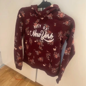 Vinröd hoodie med blommor från H&M strl 170 - Snygg vinröd hoodie från H&M med blommigt mönster och texten 'City of New York' framtill. Tröjan har huva, magficka och långa ärmar. Perfekt för dig som gillar en chill och trendig stil med lite extra färg och mönster.