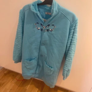 Blå teddyjacka med broderi, XXL - Mysig ljusblå teddyjacka från Bswg i storlek XXL. Jackan har fluffiga ärmar och fickor, knäppning framtill och broderade blommor på bröstet. Perfekt för dig som vill ha en chill och unik look med extra cozy vibes.