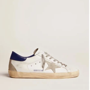 Vita sneakers Golden Goose Superstar - Golden Goose Superstar sneakers i vitt skinn med grå mockastjärna på sidan, grå snörning och blå detalj på hälen. Klassisk rund tå och platt sula. Snygg och avslappnad stil med coola detaljer som perforeringar på plösen och GGDB-text på sidan.
