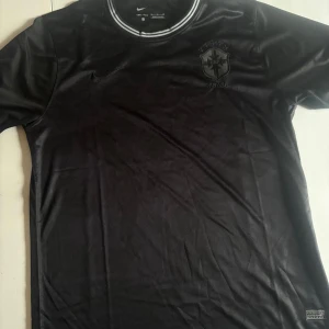 Svart Brasil Nike fotbollströja - Snygg svart Brasil fotbollströja från Nike med broderat CBF-märke och fem stjärnor på bröstet. Tröjan har rund hals med vita och grå ränder, korta ärmar och är tillverkad i Dri-FIT-material som andas. Perfekt för dig som vill sticka ut på planen eller läktaren. Kvaliten är helt ok, storleken passar för dig som har L