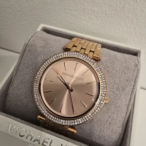 Guldig klocka från Michael Kors - Stilren guldig klocka från Michael Kors med rund urtavla i roséguld och gnistrande stenar runt boetten. Länkarmband i metall och minimalistisk design utan siffror, perfekt för dig som gillar en lyxig och elegant look. Behöver nytt batteri 