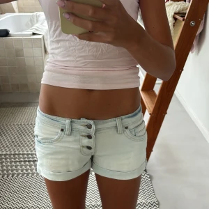 Ljusblå jeansshorts med knappar - Snygga ljusblå jeansshorts med låg midja och tre synliga knappar framtill. Klassisk femficksmodell, uppvikta benslut och cool tvättad look. Perfekta för varma dagar och enkel att matcha med allt. (Kan tänka mig att gå ner i pris vid snabb affär)