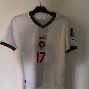 Marocko Boufal 17 fotbollströja Puma - Snygg Marocko landslagströja från Puma med Boufal och nummer 17. Vit bas med röda och gröna detaljer på krage och ärmslut, samt tryck i rött och grönt. Korta ärmar och officiella VM-märken på ärmarna. Tillverkad i lätt och ventilerande material.