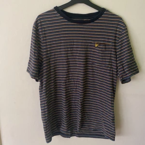 Randig t-shirt från Lyle & Scott XL - Snygg randig bommuls t-shirt från Lyle & Scott i blått, beige/orange och marinblå. Klassisk passform med korta ärmar, rund hals och bröstficka med den ikoniska gula fågeln. Perfekt för dig som gillar stilrena och bekväma plagg.